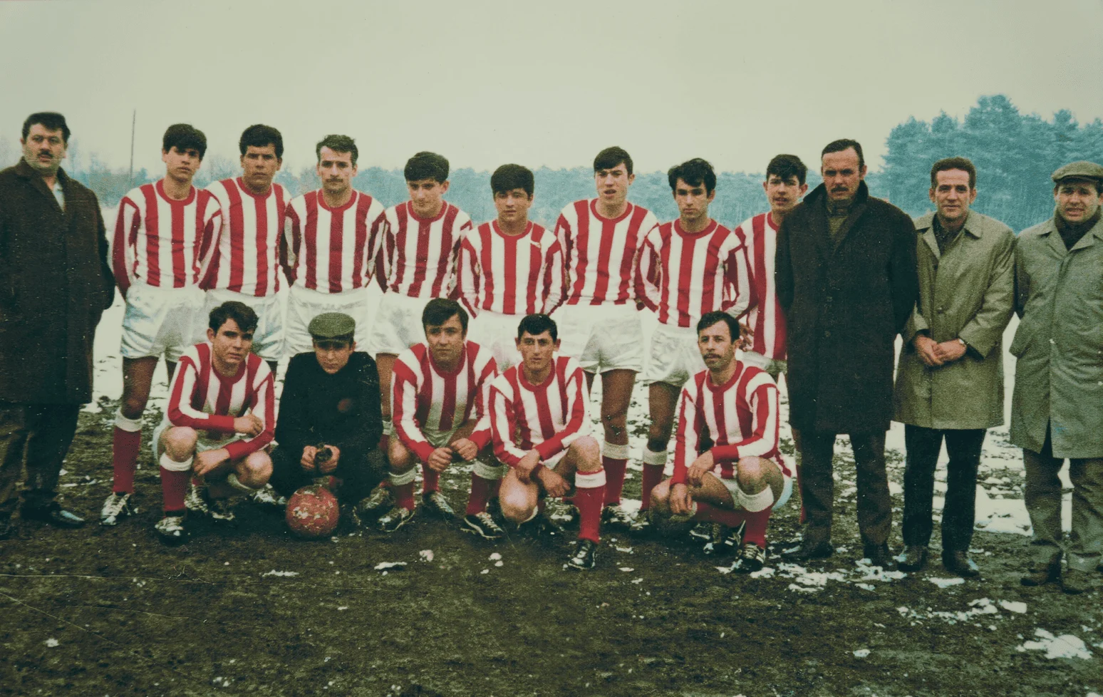 Ploegfoto van de eerste spelers van FC Anadol in rood-wit gestreepte shirts, met bestuursleden in wintermantels ernaast, op een besneeuwd veld